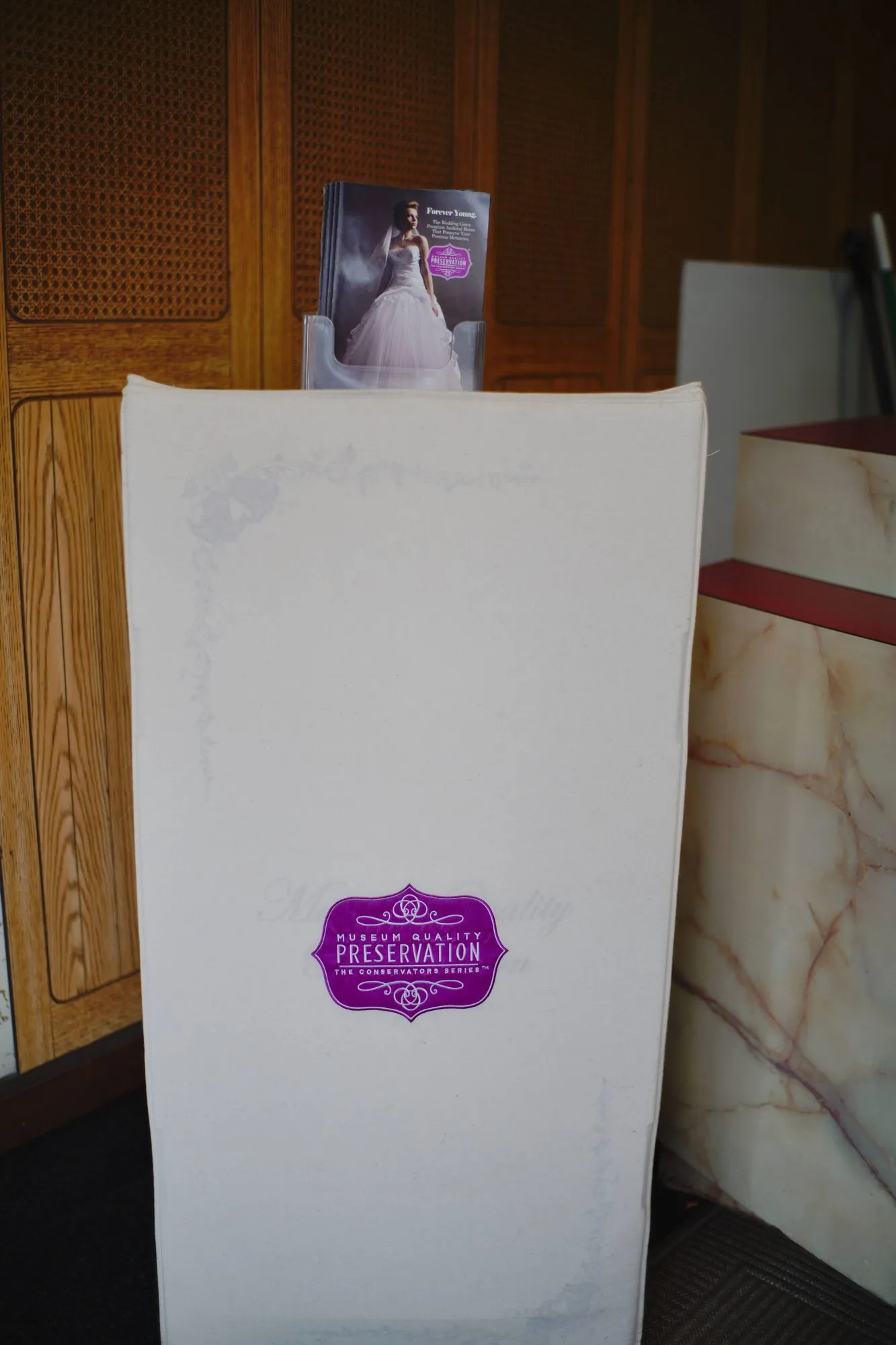 A tall wedding gown preservation box standing at the counter, labelled Museum Quality Preservation, The Conservators Series, with a gown preservation brochure behind it.